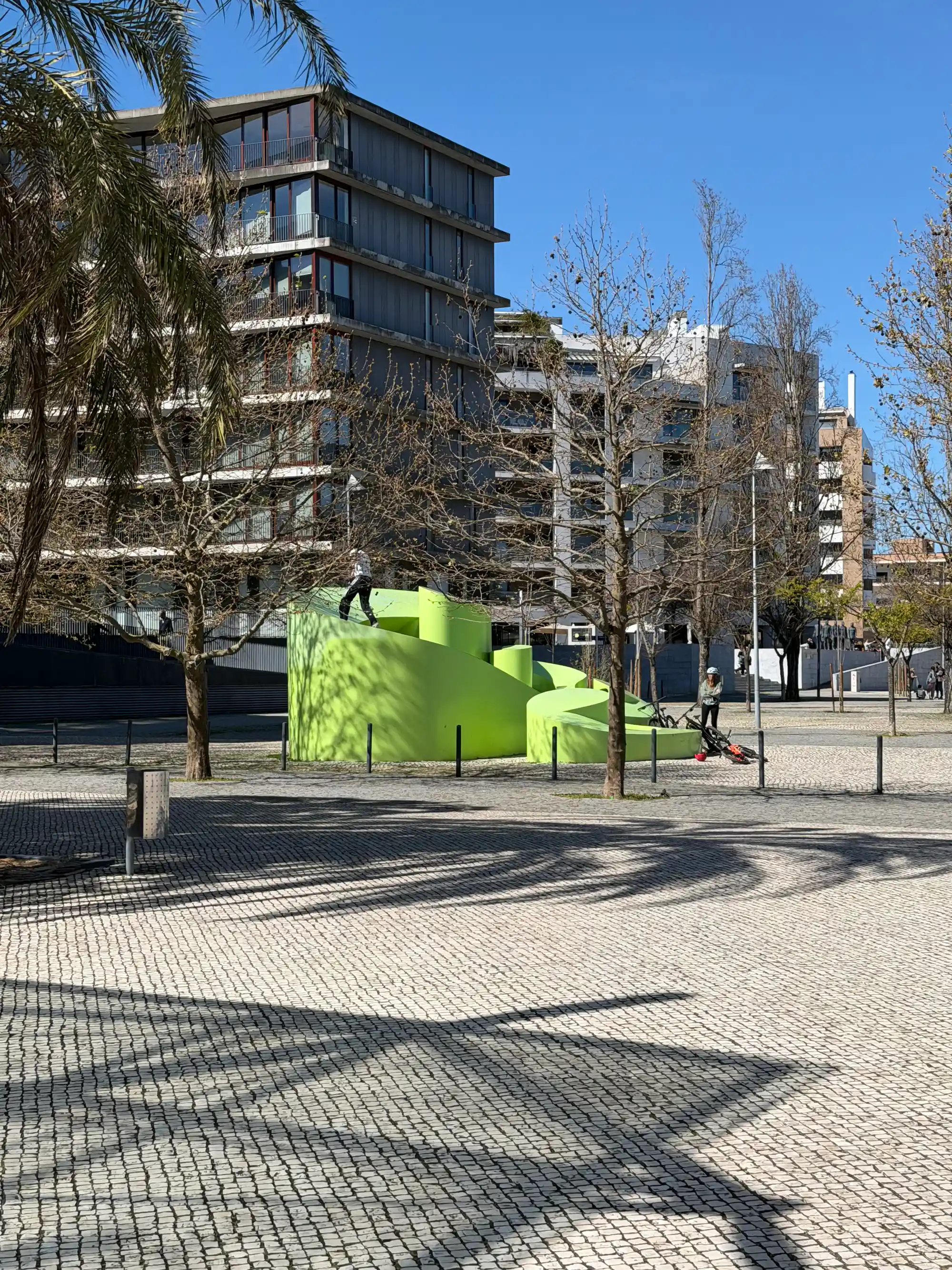 Modern green playground sculpture in Parque das Nações, Lisbon, with contemporary residential buildings and palm tree shadows on a sunny day.