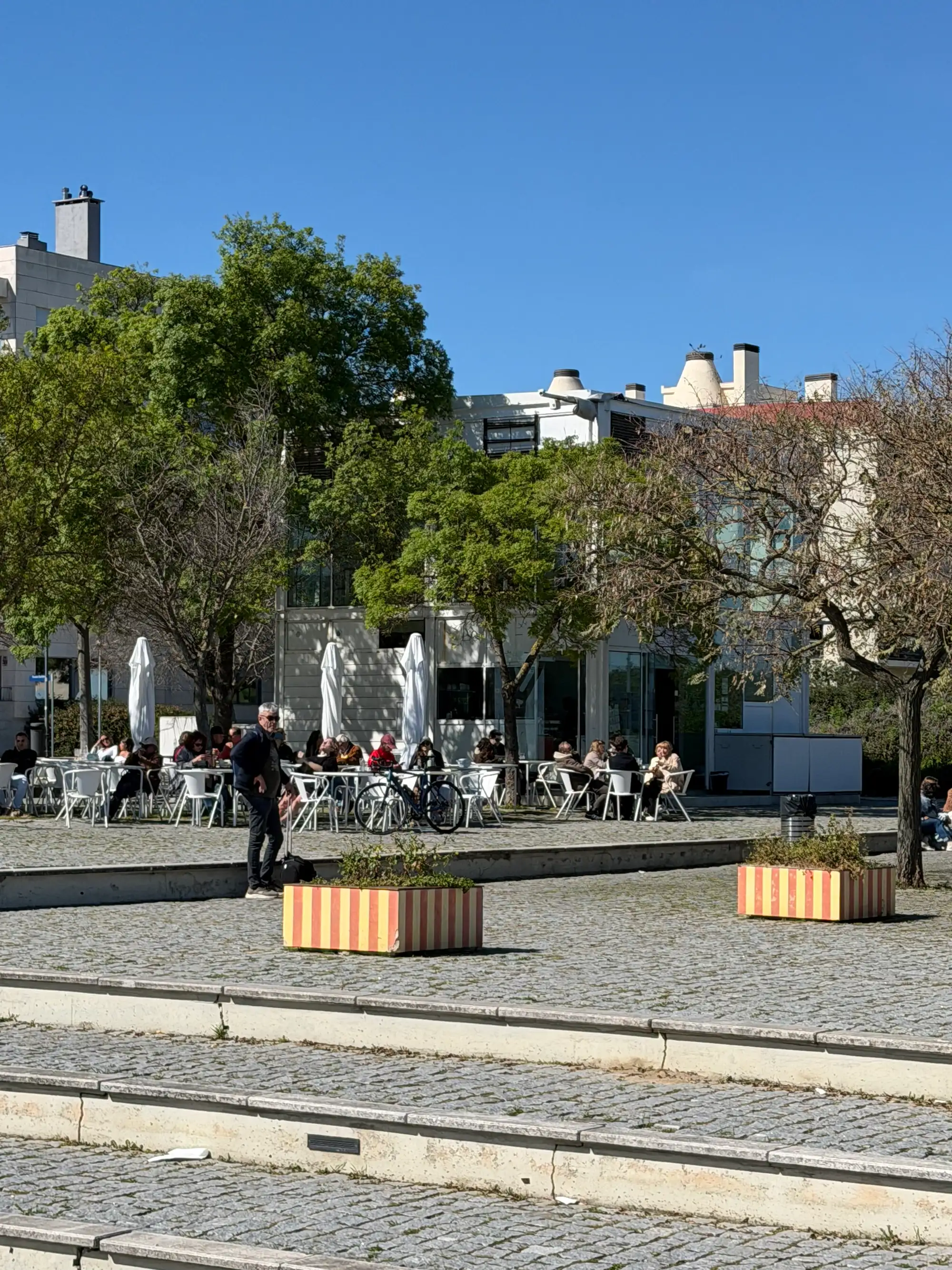 Outdoor cafe terrace with people sitting at tables in Parque das Nações, Lisbon, surrounded by trees and modern buildings on a sunny day.