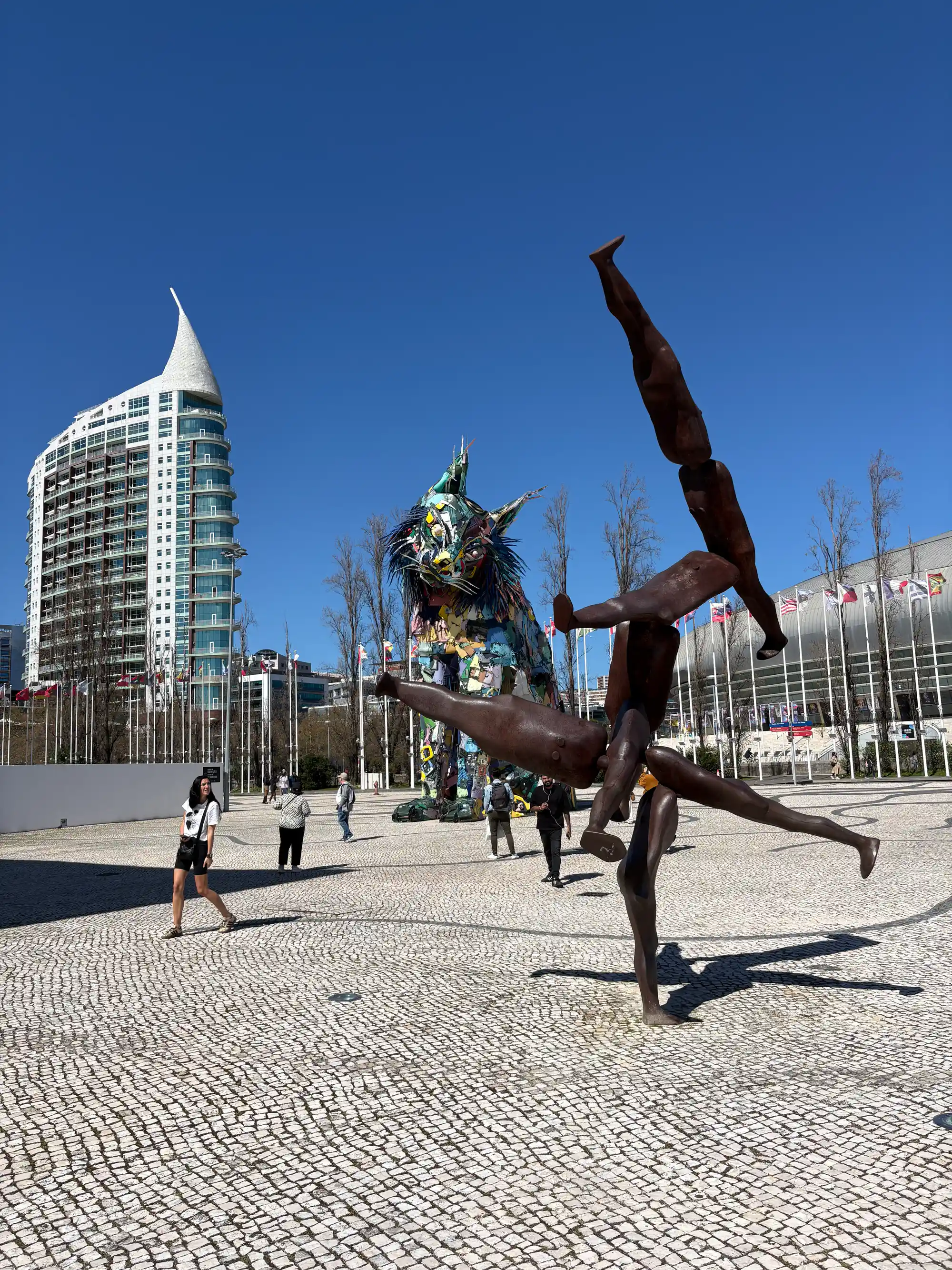 Metal sculpture of an acrobat balancing on one hand in a square at Parque das Nações in Lisbon, with a colorful lynx sculpture made of recycled materials, flags, and a modern building in the background