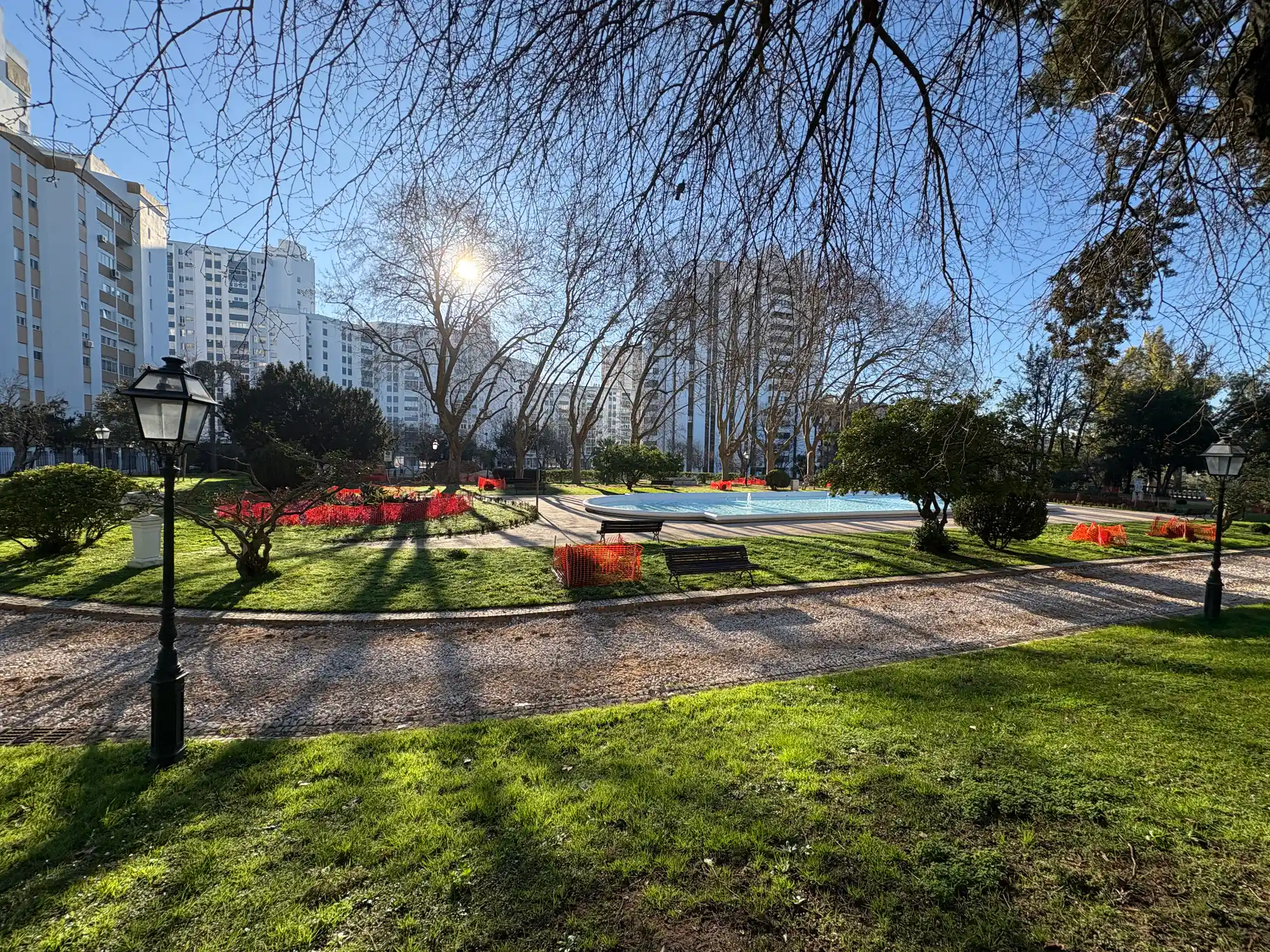 Santa Clara Lisbon park with fountain, green lawn, trees, benches, and residential buildings in the background